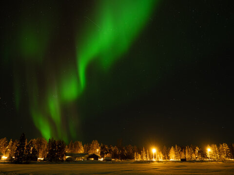 Bright green aurora borealis northern lights glowing in the night sky over a snow-covered forest and illuminated winter village in Lapland