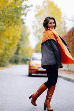 Woman walking along a quiet autumn road, wearing an orange scarf, gray coat and brown leather boots beside a car