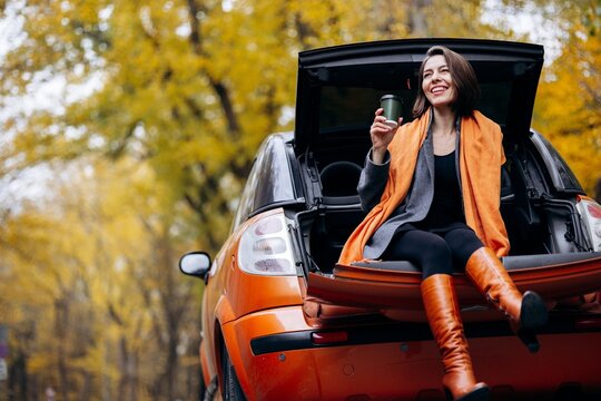Woman relaxing in the open trunk of an orange car, holding a travel mug and enjoying a peaceful break among golden autumn trees