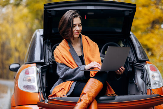 Woman seated in an open car trunk, working on a laptop during an autumn road trip amid golden foliage and woodland