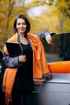 Businesswoman holding a tablet and leaning against a car, taking a confident work break outdoors among golden autumn trees