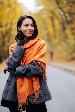 Woman wearing a vivid orange scarf and gray coat, posing on a quiet road beneath golden autumn trees