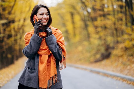 Smiling woman talking on a smartphone along a quiet road, wrapped in an orange scarf amid golden autumn foliage