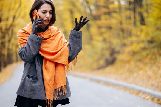 Woman talking on a phone and gesturing with frustration on a quiet road lined with golden autumn trees, wearing a scarf and gloves
