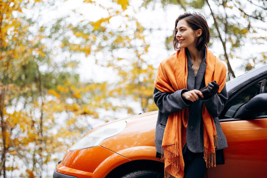 Woman in a gray coat and orange scarf, holding gloves beside an orange car beneath golden autumn foliage