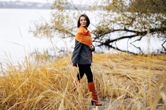Woman in an orange scarf and charcoal coat standing among golden reeds beside a lake, enjoying the stillness of an autumn day