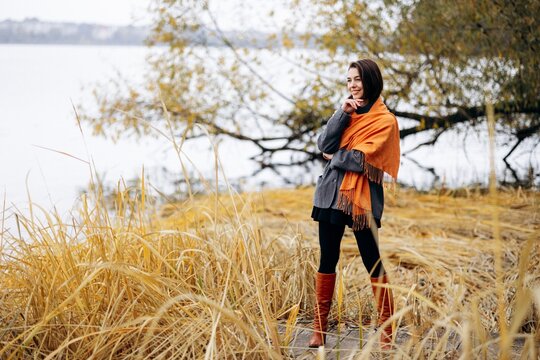 Woman wearing an orange scarf and gray coat, standing among golden reeds beside a quiet lake in autumn