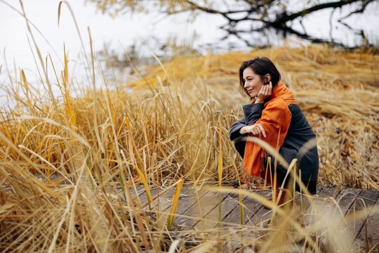Woman in an orange scarf sitting on a wooden boardwalk among golden reeds, enjoying a quiet autumn moment beside the water