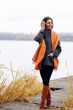 Woman standing on a lakeside dock, touching her hair in a gray coat, orange scarf, and brown boots on a calm autumn day