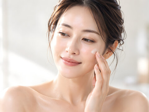 柔らかな光に包まれた白い背景で頬にクリームをなじませる女性のスキンケア美容写真 艶やかな美肌と上品なナチュラルメイクが映える自然な微笑み
