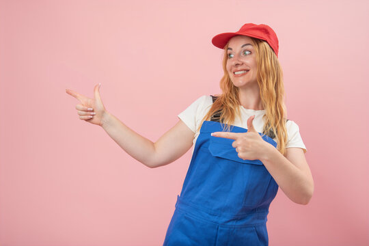 Smiling woman wearing a red cap and blue worker overalls, actively pointing fingers to the side, communicating guidance and promotion against a pink background