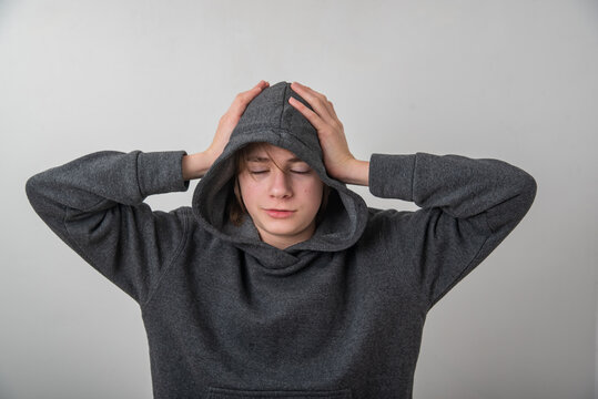 Young person wearing a gray hoodie with the hood up, eyes closed, hands on head, expressing frustration, fatigue, or a headache, representing common challenges of youth