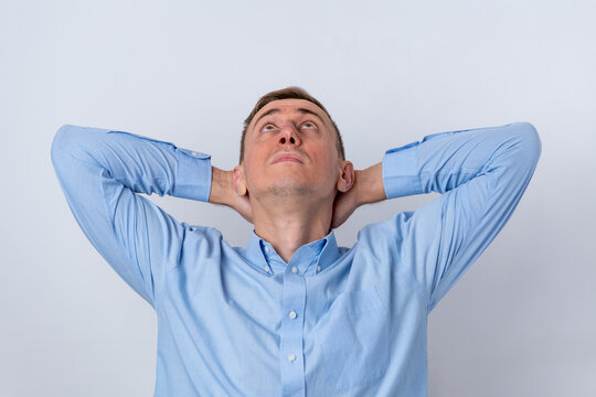 Young man wearing a blue shirt, resting with hands behind his head on a white background, contemplating new ideas and opportunities with an upward gaze