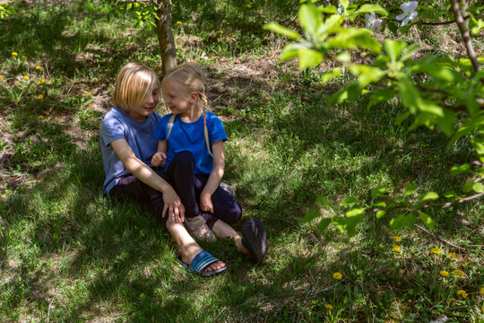 Two children sitting on grass under trees sharing quiet conversation and smiling together while one rests hand on other's knee, sunlit leafy outdoor setting, candid moment of friendship and childhood