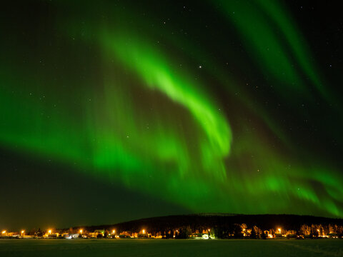 Green aurora borealis northern lights glowing in the night sky over a snow-covered forest and illuminated winter village in Lapland