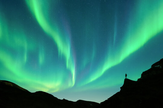Vivid green northern lights sweeping across a starry night sky above a dark mountain ridge with tourist silhouette. Dramatic natural aurora display with flowing curtains of light