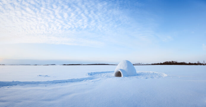 Panorama with small snow igloo standing alone on a vast frozen plain beneath a pale blue sky filled with delicate clouds. Minimalist winter landscape with soft light and a distant dark horizon