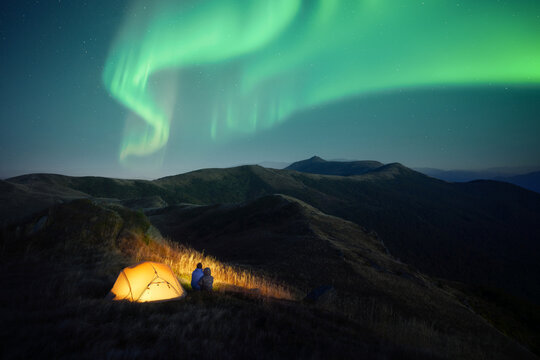 Green northern lights illuminate a dark mountain landscape with a glowing tent beneath the night sky. Two hikers near his tent and distant stars create a peaceful camping scene