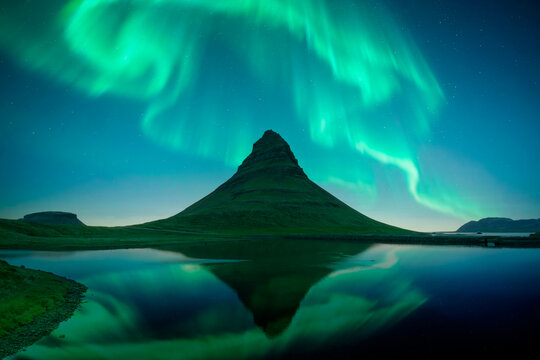 A cone-shaped Kirkjufell mountain reflects in a still lake beneath vivid green northern lights. Dramatic Icelandic landscape. Aurora borealis. Polar lights. Iceland