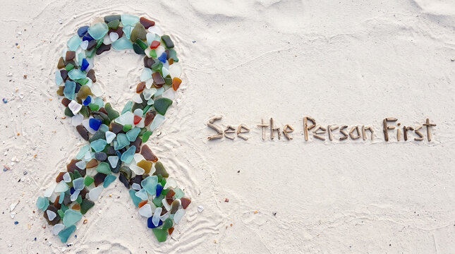 A awareness ribbon made of colorful sea glass on white sand with text see the person first with formed various pieces glass arranged sandy sandy beach next ribbon shape understanding