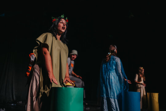 A female actress stands at the front of the stage performing in costume. Other cast members appear behind her in a theatrical ensemble scene.