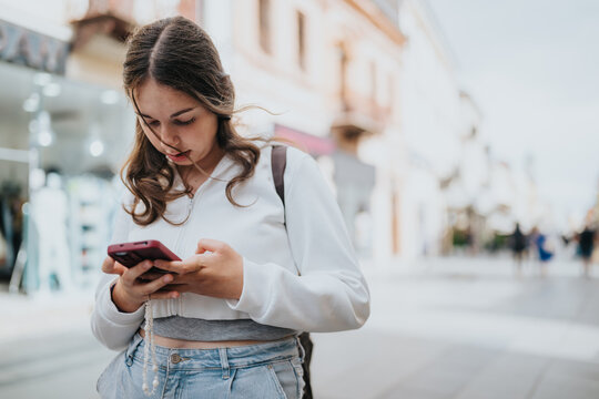 A teenage girl wearing a white cropped jacket and jeans looks at her smartphone on a city shopping street. She walks along a pedestrian street checking her phone in a casual everyday moment.