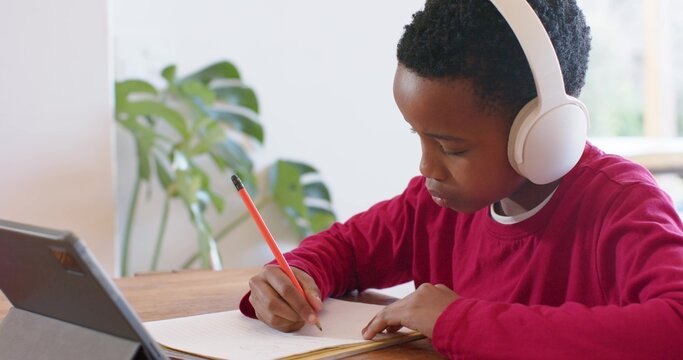African American male child writing in notebook at wood table by window, red shirt, orange pencil