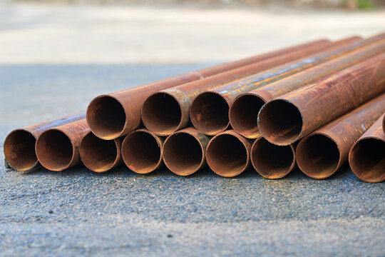 Stack of rusty metal pipes lying on asphalt ground, pile of steel tubes arranged in row, replacement work. Stack of weathered iron pipes stored outdoors on paved surface. Construction materials