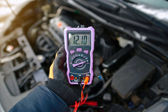 Car battery test with digital multimeter displaying 12.10 volts, electrical diagnostics on car battery inside an open engine bay in cold winter conditions. Low voltage, weak, discharged battery issues