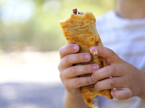 A child holds a bitten sausage roll in both hands, with flaky pastry around the filling, a bakery snack that connects savory convenience food with children's eating habits and quick meals on the go.