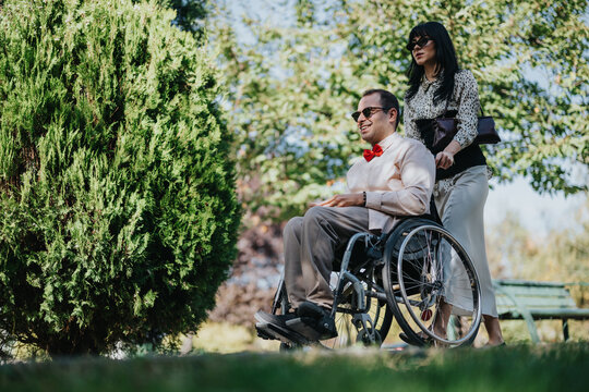 Man uses a wheelchair while a woman provides caregiver support through a sunny park, showing disability inclusion, mobility and assistance.