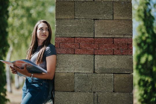 College student holds a book beside a brick pillar on campus, representing education, independent study and young adult life.