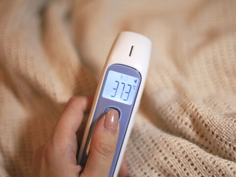 Hand holds a digital thermometer reading 37.3 degrees Celsius beside a knitted blanket, with the result providing a body temperature measurement for home healthcare and personal fever monitoring.