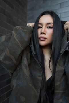 Close-up portrait of a beautiful Asian woman holding the hood of a camouflage jacket outdoors. Looking at camera on a dark brick wall background. Stylish urban street fashion look. Pretty glam girl