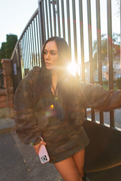 Stylish young Asian woman in a camouflage jacket posing near a metal fence during sunset, holding a smartphone in warm sunlight with lens flare