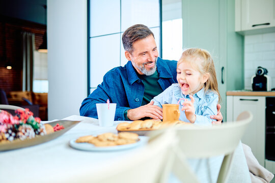 Father and young daughter sharing cookies at the kitchen table, laughing together during a joyful family snack
