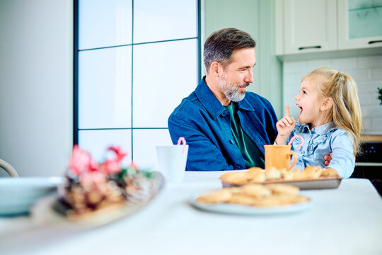 Father and young daughter sharing a lively conversation at the kitchen table, with cookies and mugs set before them