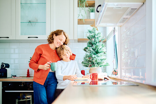 Mother and son sharing a tender holiday moment, preparing treats together in a bright kitchen beside a Christmas tree