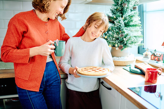 Mother and son smiling together, the boy carrying a freshly baked pie beside a decorated Christmas tree in their kitchen