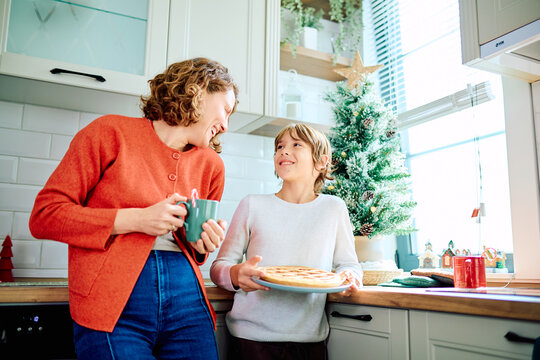 Mother and son smiling together in a bright kitchen, sharing a freshly baked pie beside a decorated Christmas tree