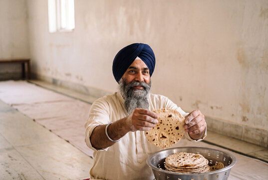 Smiling senior South Asian man in navy turban serving fresh roti in community kitchen, langar seva concept with copy space