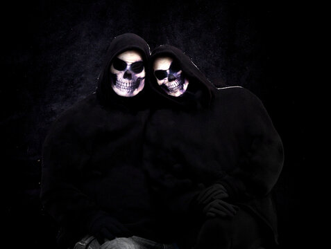 Two people in skull masks and black clothes against a black background.