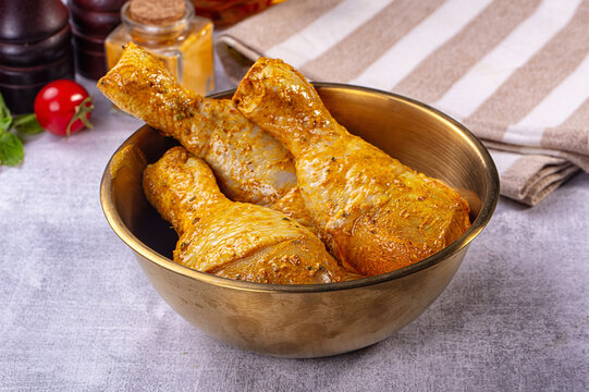 Brass bowl with striped kitchen towel holding three raw chicken drumsticks coated in yellow spice marinade, cherry tomato and spice jars nearby, marinated poultry