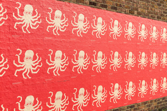 Repeating pattern of white octopus illustrations on red posters pasted along an outdoor brick wall in perspective view. Sunlight illuminates the textured paper posters and brick building, capturing ur