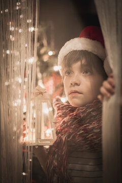 Close-up portrait of a young Boy wearing a santa hat standing by a window holding an illuminated lantern at Christmas