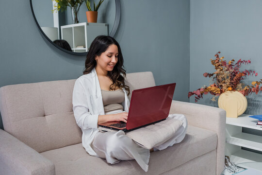 young Hispanic woman using laptop sitting on sofa at home in Latin America