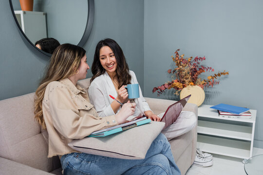 Young hispanic couple of women friends using laptop and sitting on the sofa at home in Latin America