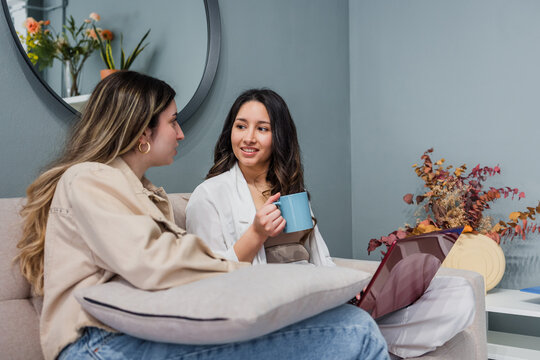 Young hispanic couple of women friends using laptop and sitting on the sofa at home in Latin America