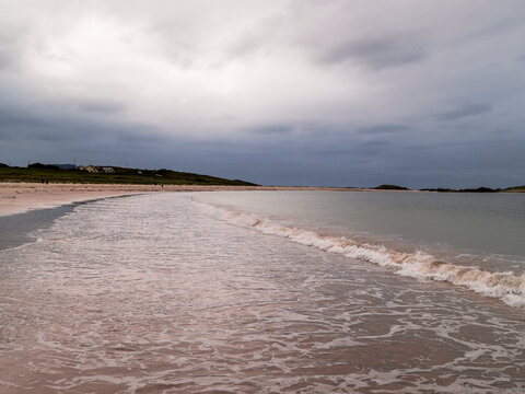 Pink-hued seafoam crests near a mossy rock on a calm beach. Waves gently roll toward shore under an overcast sky. Distant hills fade into soft mist on horizon