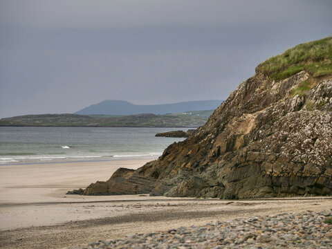 Calm coastal scene with rocky cliffs and sandy beach. Gray skies hover over distant hills and gentle waves. Nature's quiet strength shown through rugged terrain and soft shoreline